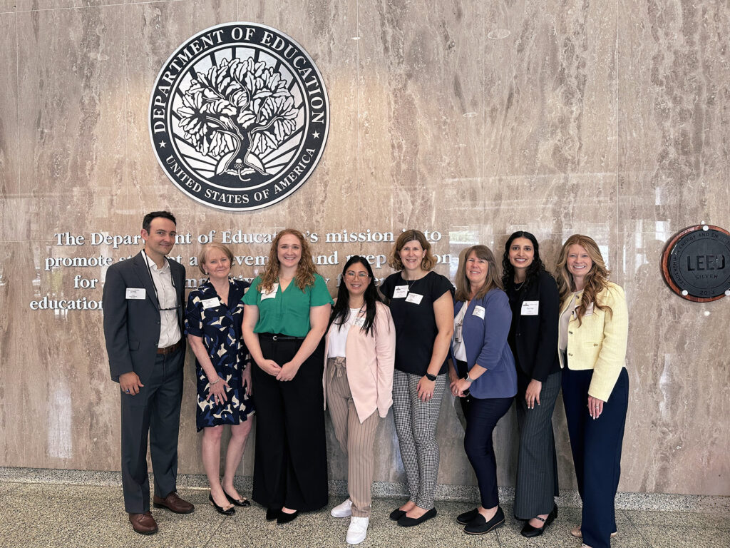 A group of eight people are standing in front of the Department of Education emblem and mission statement on a marble wall. They are dressed in professional attire, with name badges visible on their clothing. From left to right, there are three men and five women. The individuals are smiling and posing for the photo, suggesting they are attending an event or meeting at the Department of Education in the United States. The wall behind them also has a LEED Silver certification plaque.