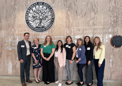 A group of eight people are standing in front of the Department of Education emblem and mission statement on a marble wall. They are dressed in professional attire, with name badges visible on their clothing. From left to right, there are three men and five women. The individuals are smiling and posing for the photo, suggesting they are attending an event or meeting at the Department of Education in the United States. The wall behind them also has a LEED Silver certification plaque.