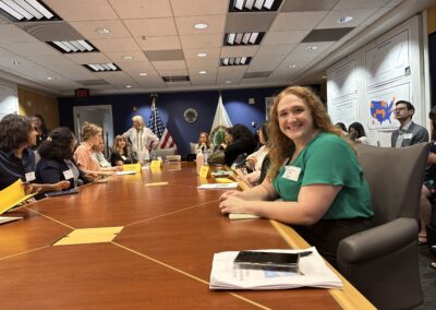 A woman in a green blouse is sitting at a large conference table, smiling at the camera. The table is surrounded by other attendees engaged in discussions, with name cards, papers, and folders spread out. The background features flags and the Department of Education seal on the wall.