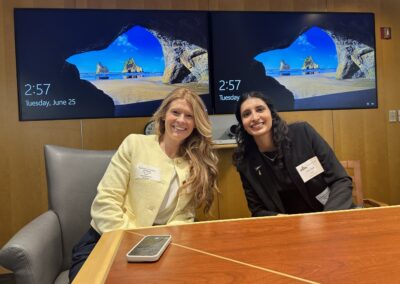 Two women are seated at a conference table, smiling at the camera. The woman on the left is wearing a light yellow blazer, and the woman on the right is wearing a black blazer. Behind them, two large monitors display a scenic beach image and the time, 2:57, on Tuesday, June 25.