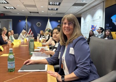 A woman in glasses wearing a blue blazer is seated at a conference table, smiling at the camera. She has a tablet and a bottle of Pellegrino water in front of her. Other attendees are engaged in discussions around the table, with flags and the Department of Education seal visible in the background.