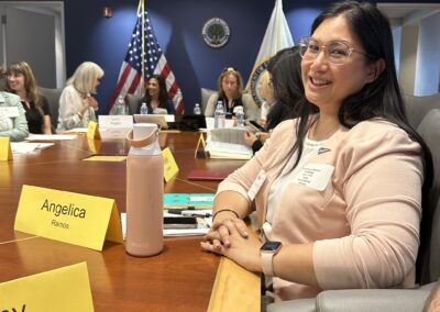 A woman in glasses wearing a peach cardigan is sitting at a conference table, smiling at the camera. Her name card is in front of her, along with a water bottle and documents. Other attendees are seated around the table, with flags and the Department of Education seal in the background.