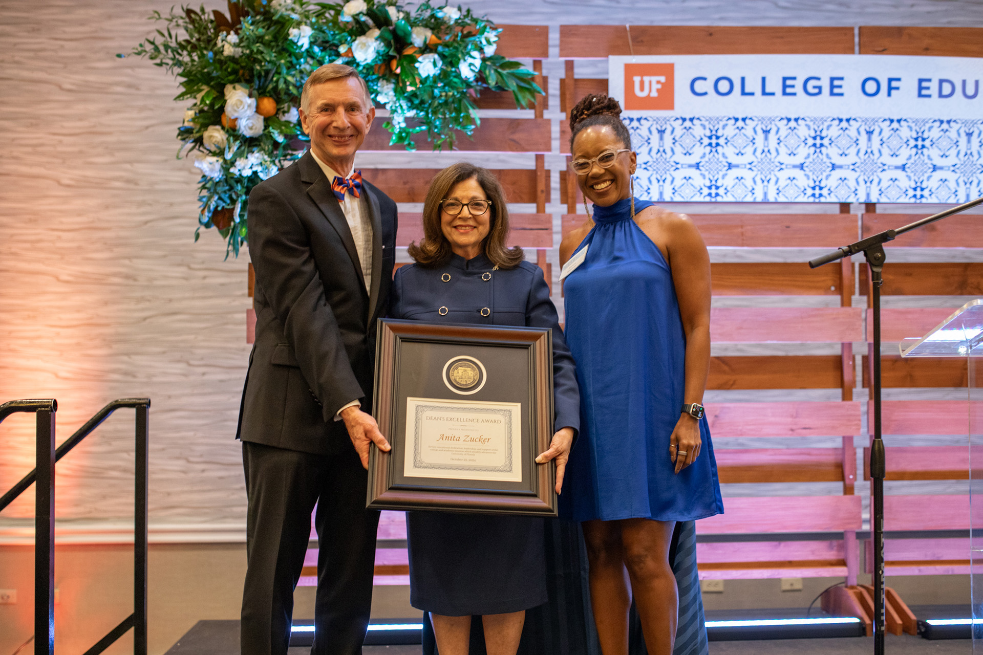 Three individuals stand on a stage beside a microphone, holding a framed award plaque. The person in the center holds the plaque, which reads "Dean’s Excellence Award." They are dressed in formal attire, surrounded by a backdrop with a wooden structure, floral arrangement, and a partial sign that reads "UF College of Education" with a blue decorative pattern.