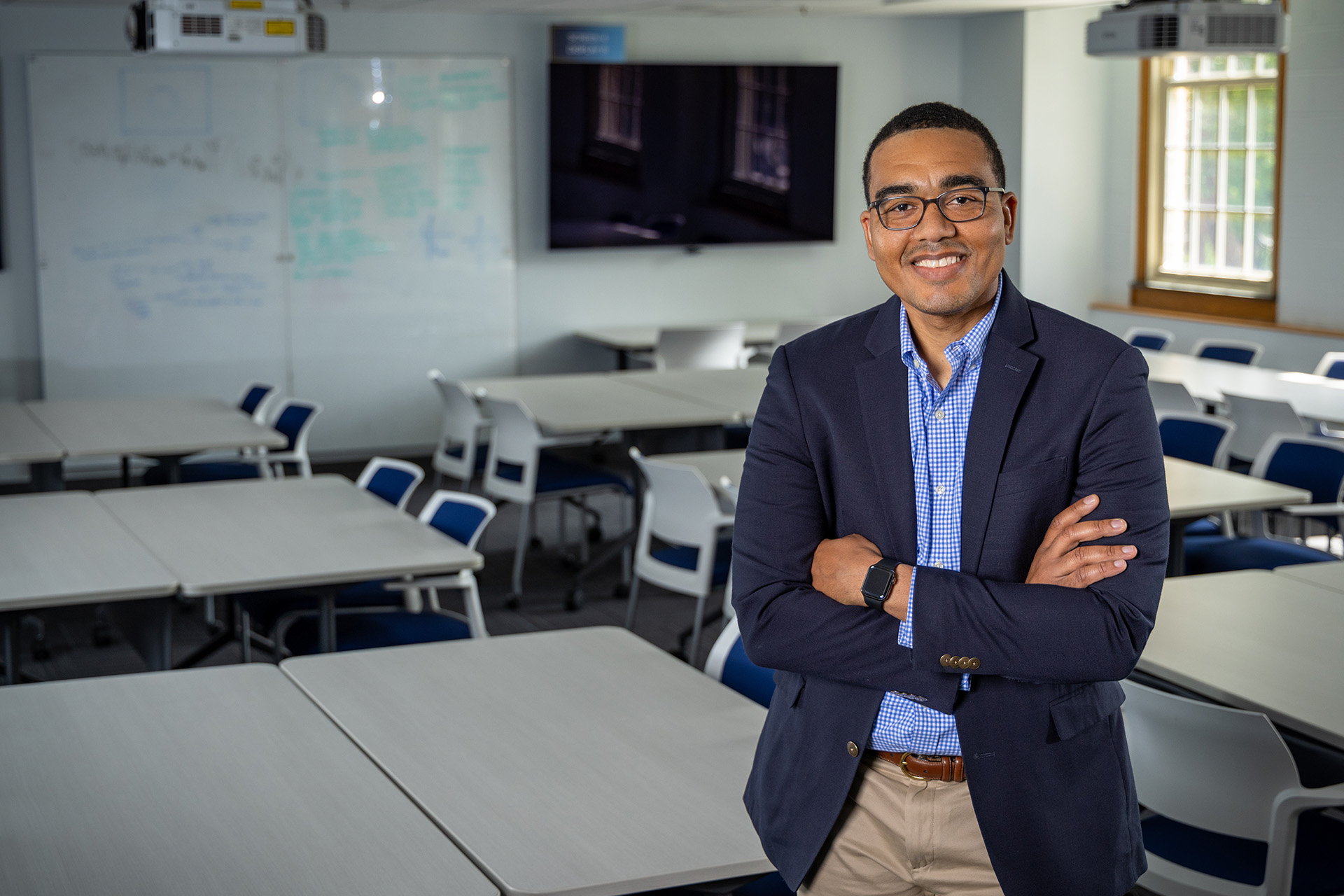 A man wearing a navy blazer over a blue checkered shirt stands with arms crossed in a modern classroom. The room is equipped with white rectangular tables and blue chairs, a large whiteboard with diagrams and writing, a flat-screen TV mounted on the wall, a ceiling-mounted projector, and large windows that allow natural light to enter.