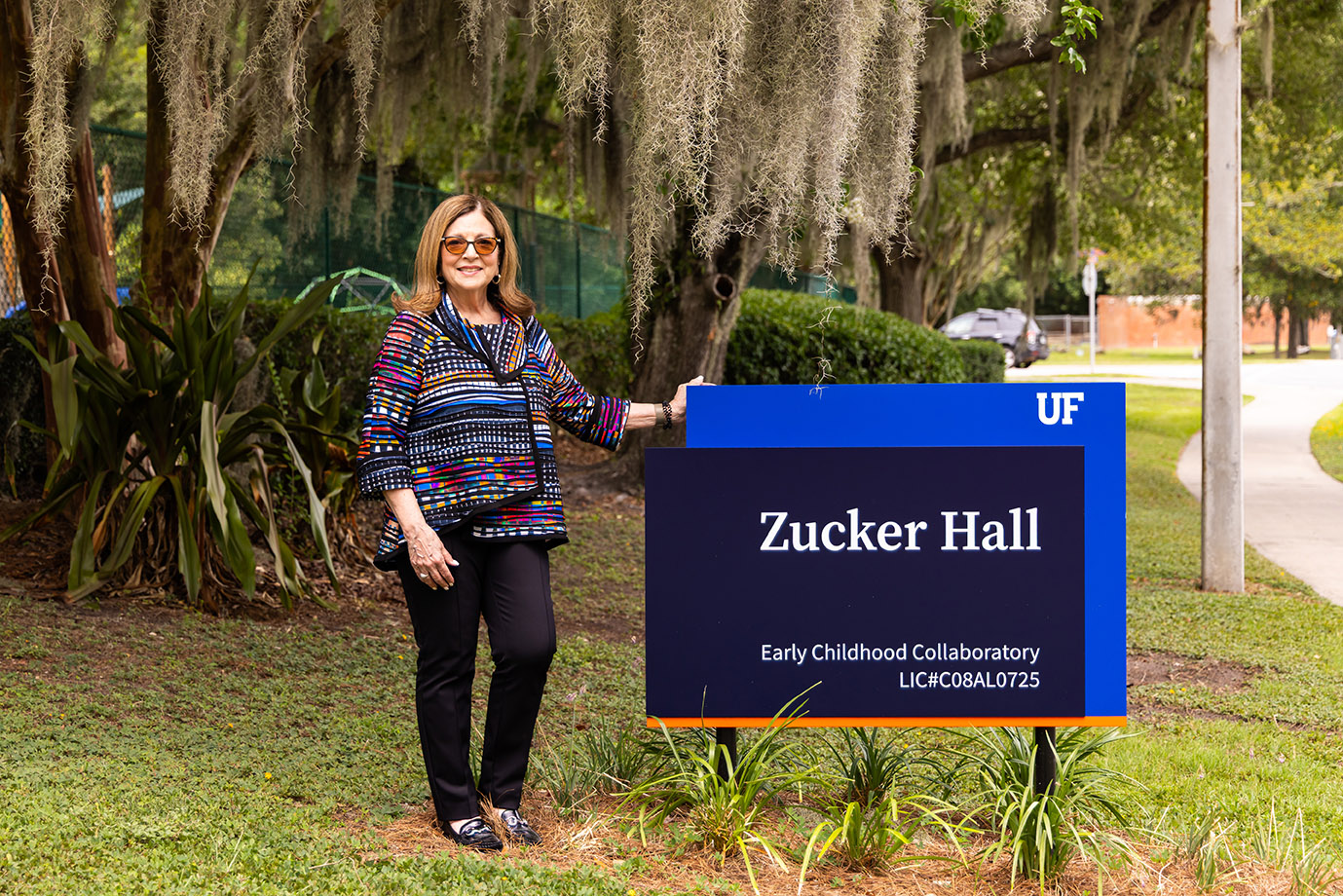 Anita Zucker sign A smiling woman standing beside a dark blue and orange sign that reads "Zucker Hall, Early Childhood Collaboratory, LIC# C08AL0725", with the University of Florida logo in the top right corner. The backdrop consists of greenery with a green net fence and trees with Spanish moss.