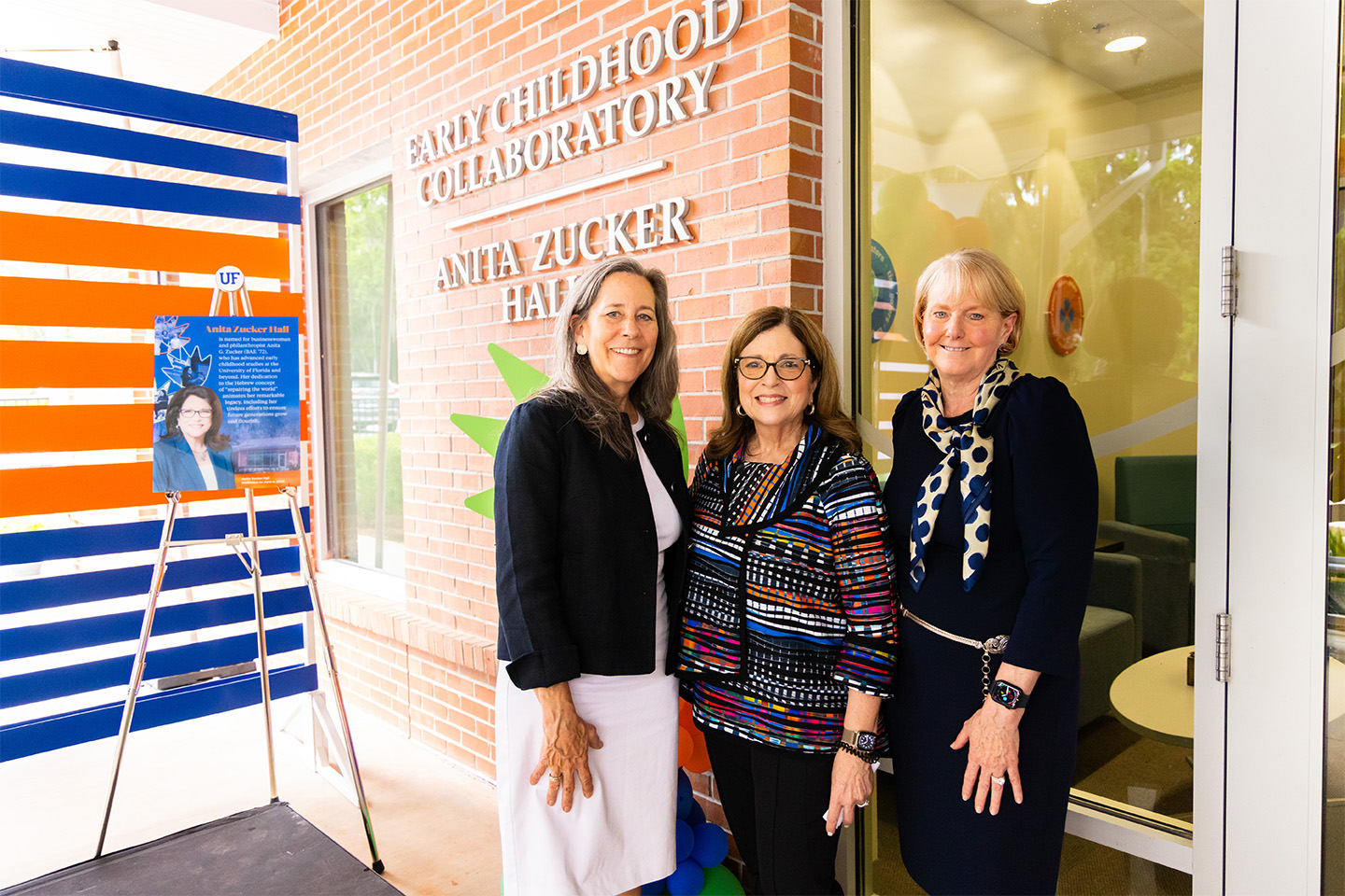 Maureen Conroy Anita Zucker Pat Snyder - landscape Three women smile while posing for a photo in front of the brick exterior of a building. Silver lettering on the wall reads, "Early Childhood Collaboratory, Anita Zucker Hall." To their left is a sign on an easel next to a blue and orange backdrop.